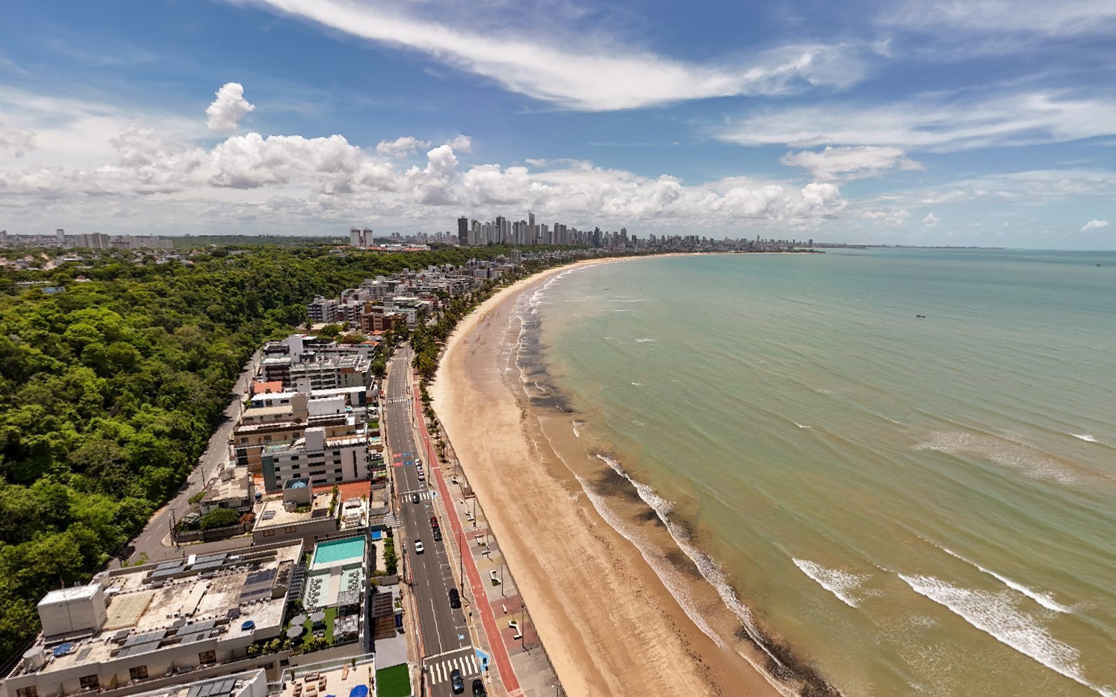 Vista aérea da enseada do Cabo Branco, com a curva da praia, a mata da falésia à esquerda e os prédios de João Pessoa no horizonte