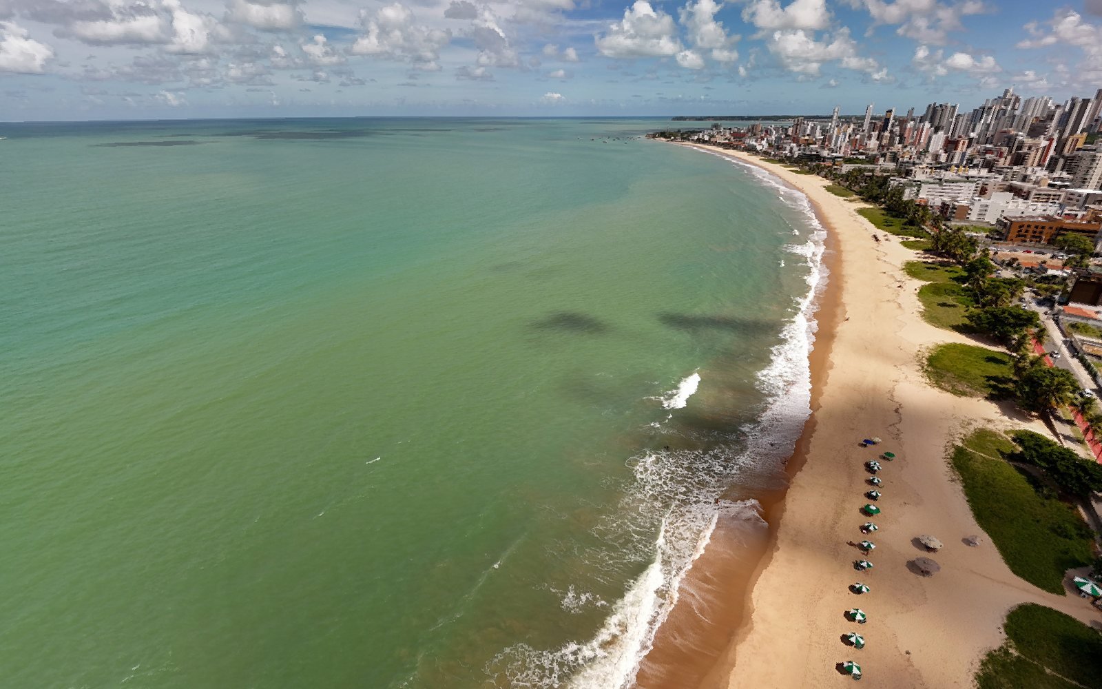 Vista aérea da orla de Manaíra, em João Pessoa, com o mar verde à esquerda, a areia com guarda-sóis e os prédios do bairro à direita